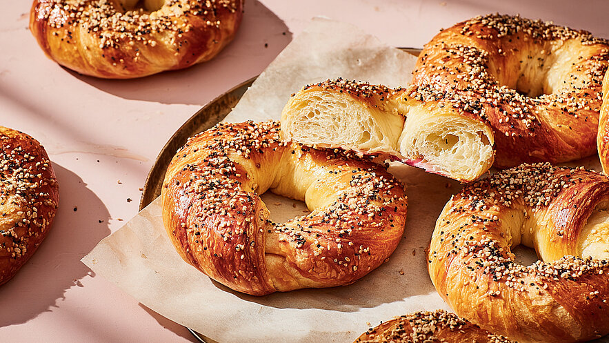 Eine Auswahl frisch gebackener Bagels, goldbraun und mit einer Mischung aus Sesam- und Schwarzkümmelsamen bestreut. Einer der Bagels ist zur Hälfte geschnitten, um die luftige, weiche Textur im Inneren zu zeigen. Hintergrund in sanften, neutralen Farbtönen.
