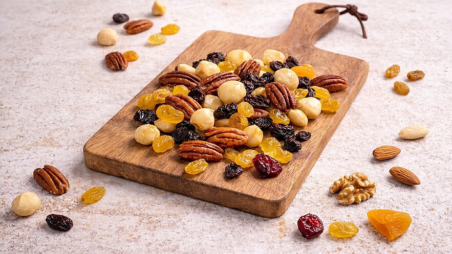 Eine Holzplatte ist mit einer bunten Mischung aus Nüssen und Trockenfrüchten belegt. Darunter befinden sich Pekannüsse, Macadamianüsse, Rosinen, sowie getrocknete Aprikosen und andere Früchte. Die Gesamtanordnung vermittelt einen Eindruck von frischen, gesunden Snacks.