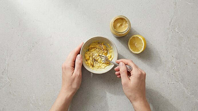 Zwei Hände halten eine Schale und rühren mit einer Gabel eine gelbe Paste; daneben liegen eine halbe Zitrone und ein kleines Glas mit gelbem Aufstrich.