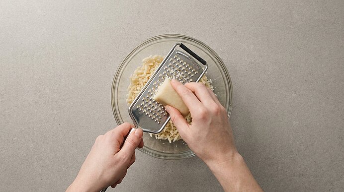 Nahaufnahme: Hände reiben ein Käsestück über eine Metallreibe, geriebener Käse fällt in eine Glasschüssel.