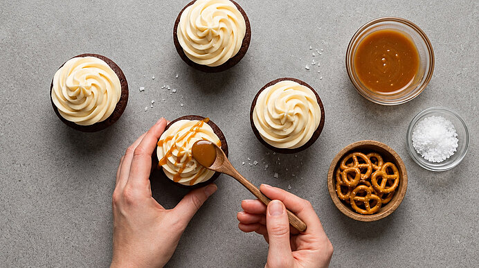 Zwei Hände garnieren Cupcakes mit Karamell und Salzbrezeln. Daneben stehen Schälchen mit Karamell, Brezeln und Salz.