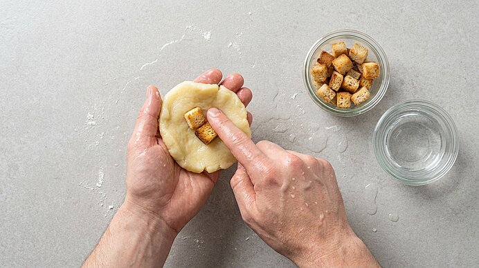 Zwei Hände formen einen runden Teigling; mit dem Zeigefinger wird ein Brotwürfel in die Mitte gedrückt. Daneben eine Schale mit weiteren Brotwürfeln und ein Glas Wasser.