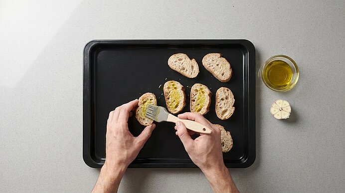 Zwei Hände bestreichen Baguettescheiben auf einem schwarzen Backblech mit Pinsel und Olivenöl.