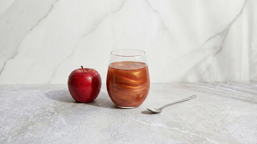 Ein roter Apfel liegt neben einem Glas mit einer braunen, schimmernden Flüssigkeit. Ein kleiner Löffel befindet sich neben dem Glas. Der Hintergrund ist hell und marmoriert.