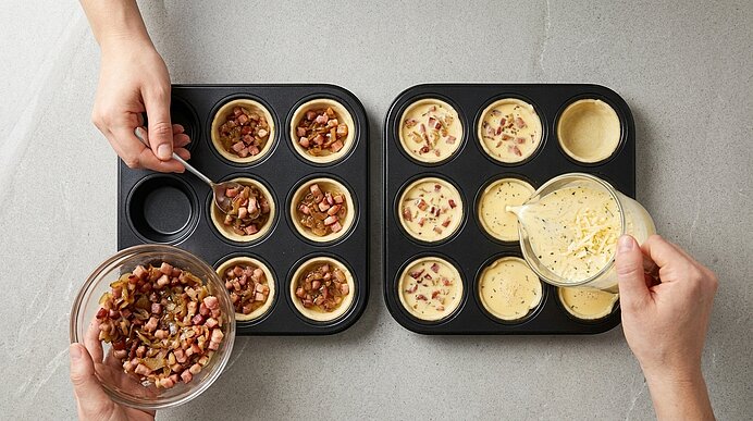 Zwei Hände befüllen Teigförmchen in Muffinblechen mit Speck-Zwiebel-Mischung und gießen Eiermasse hinein.