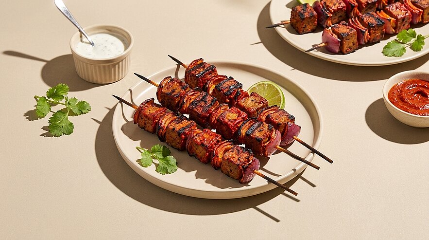 Drei gegrillte Spieße mit mariniertem Fleisch und Gemüse auf einem Teller, garniert mit Koriander und einer Limettenscheibe; daneben Schälchen mit weißem Joghurt-Dip und roter Chilisauce.