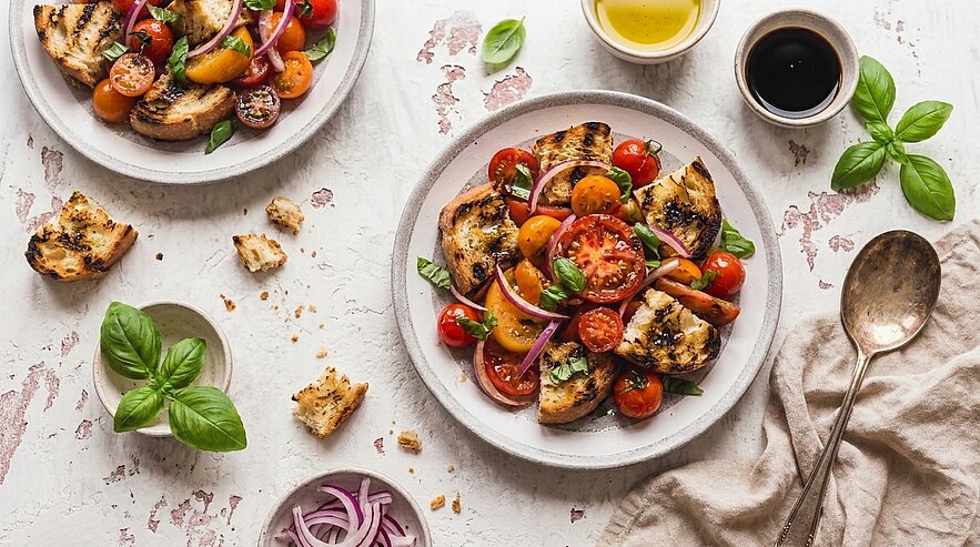 Zwei Teller mit gegrilltem Brot und einem bunten Salat aus Tomaten, roten Zwiebeln und frischem Basilikum. Olivenöl und Balsamico-Vinaigrette in kleinen Schalen neben den Tellern. Die Szene ist hell und einladend mit einer rustikalen Textur im Hintergrund.