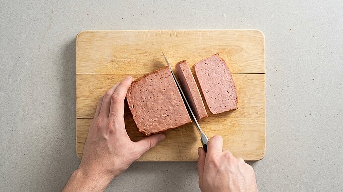 Auf einem Holzbrett schneiden zwei Hände ein Stück Fleischkäse mit einem Messer in dicke Scheiben.