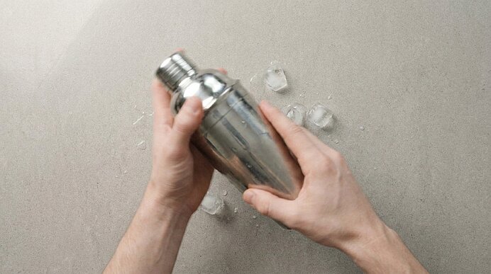Zwei Hände halten einen silbernen Cocktailshaker auf einer hellgrauen Arbeitsplatte mit vereinzelten Eiswürfeln.