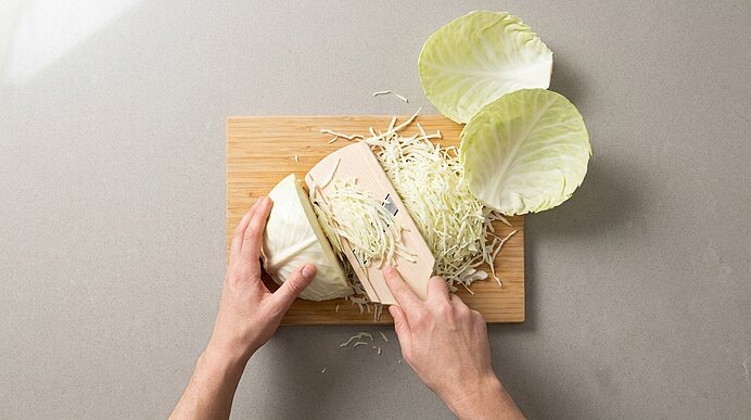 Hände raspeln Weißkohl mit einem Messer auf einem Holzbrett; daneben liegen Kohlblätter und feine Kohlstreifen.