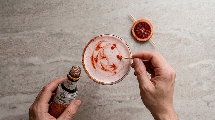 Hände verzieren einen rosa Schaumcocktail in einer Coupette, indem mit einem Fläschchen Bittertropfen Muster auf den Schaum gesetzt werden; getrocknete Blutorangenscheibe auf Spieß daneben.