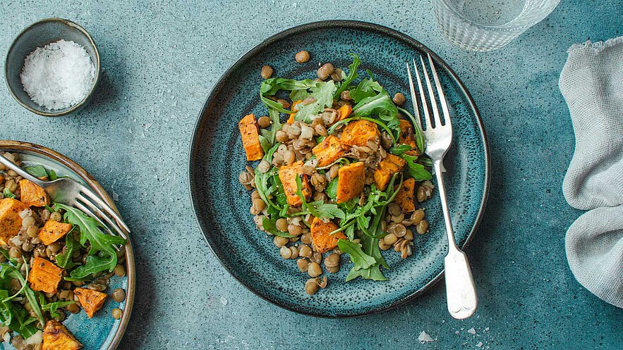 Ein Teller mit einem frisch zubereiteten Salat aus Rucola, Linsen und geröstetem Kürbis. Der Salat ist bunt und appetitlich angerichtet. Im Hintergrund ist ein kleiner Schüssel mit Salz und ein Glas mit Wasser zu sehen.
