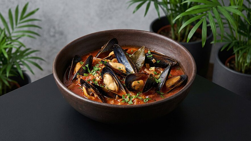 Eine dunkelbraune Schüssel enthält eine schmackhafte Muschel-Suppe, garniert mit frischen Kräutern. In der Brühe sind mehrere Muscheln sichtbar, die teils geöffnet sind. Der Hintergrund zeigt unscharfe Pflanzen in Töpfen.