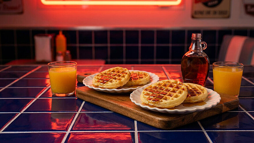 Auf einem Holzbrett liegen zwei Teller mit Waffeln, die mit Sirup überzogen sind. Daneben steht eine Glasflasche mit Ahornsirup und ein Glas mit orangefarbnem Saft. Der Tisch ist mit blauen Fliesen dekoriert.