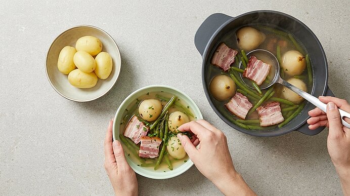 Hände schöpfen aus einem Topf eine klare Brühe mit kleinen Kartoffeln, grünen Bohnen und Speckstücken in eine Schüssel; daneben eine kleine Schale mit Kartoffeln.