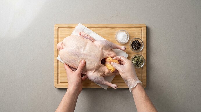 Mit Einweghandschuhen bedeckte Hände füllen eine Zitronenhälfte in ein rohes, ganzes Hähnchen auf einem Holzbrett; daneben Schälchen mit Salz, Pfeffer und Kräutern.