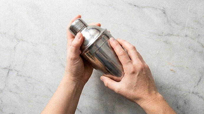 Zwei Hände halten einen silbernen Cocktail-Shaker über einer hellen Marmorarbeitsfläche, bereit zum Mixen.