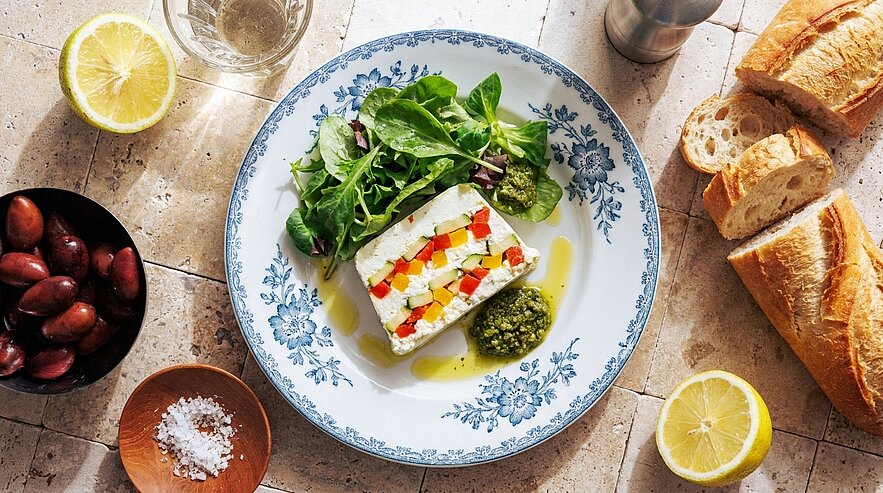 Teller mit Frischkäseterrine, belegt mit fein gewürfelten roten und gelben Paprika, serviert mit grünem Pesto, frischem Blattsalat und einem Schuss Olivenöl.