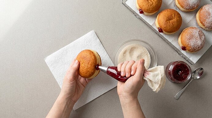 Hände füllen einen Berliner mit roter Marmelade aus einer Spritztüte; daneben Puderzucker, Marmeladenglas und ein Blech mit mehreren puderzuckerbestäubten Krapfen.