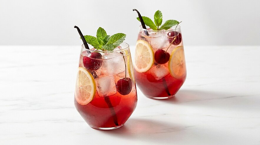 Zwei Gläser mit roten Sommerdrinks auf hellem Untergrund, gefüllt mit Eis, Zitronenscheiben, Kirschen, Minzblättern und mit Vanilleschoten garniert.