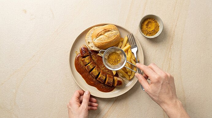 Teller mit geschnittener Currywurst in Currysauce, Pommes frites und Brötchen; Hand streut Currypulver durch ein Sieb über die Wurst.