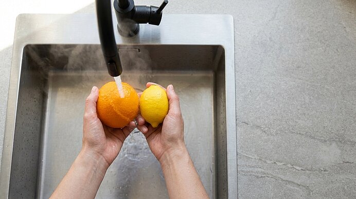 Zwei Hände halten und waschen eine Orange und eine Zitrone unter fließendem Wasser im Spülbecken.