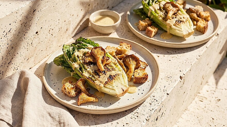 Zwei Teller mit gegrillten Romanasalat‑Hälften, gebratenen Pfifferlingen, Croutons und einem kleinen Schälchen cremigem Dressing.