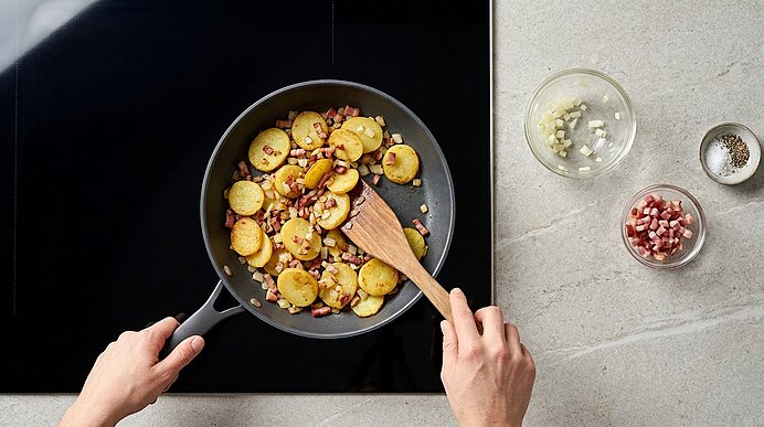 Hände rühren mit einem Holzspatel in einer Pfanne gebratene Kartoffelscheiben mit Speckwürfeln und Zwiebeln auf einem Cerankochfeld.