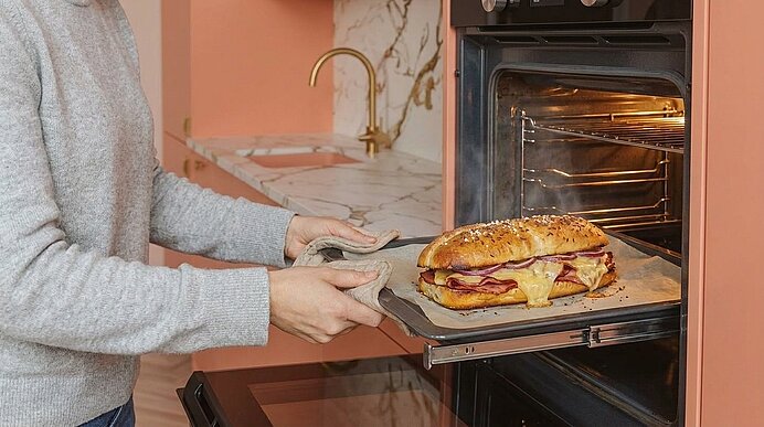 Hände mit Küchenhandschuhen nehmen ein großes, mit geschmolzenem Käse und Schinken überbackenes Sandwich auf einem Backblech aus einem eingeschalteten Ofen.