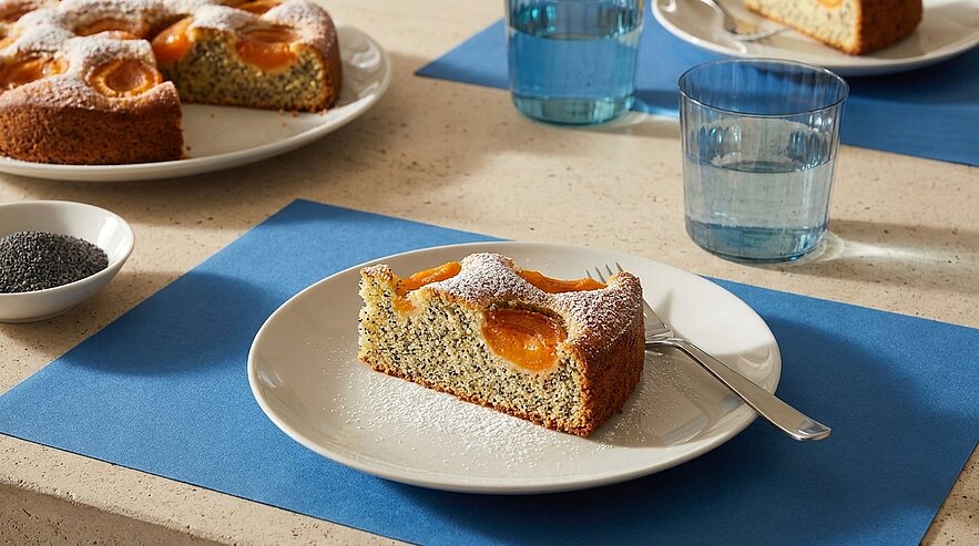 Ein Stück Mohnkuchen mit Aprikosenstücken liegt auf einem weißen Teller. Der Kuchen ist bestäubt mit Puderzucker. Daneben steht ein Glas Wasser. Im Hintergrund sind weitere Kuchen und ein kleines Schälchen mit Mohnsamen zu sehen.