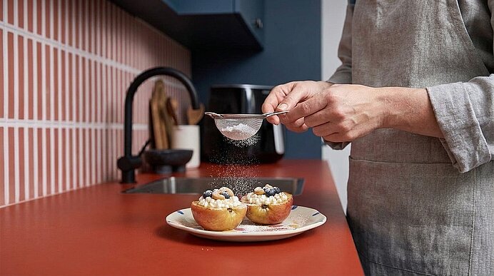 Hände streuen Puderzucker über gebackene Apfelhälften mit Creme und Beeren auf einem Teller.