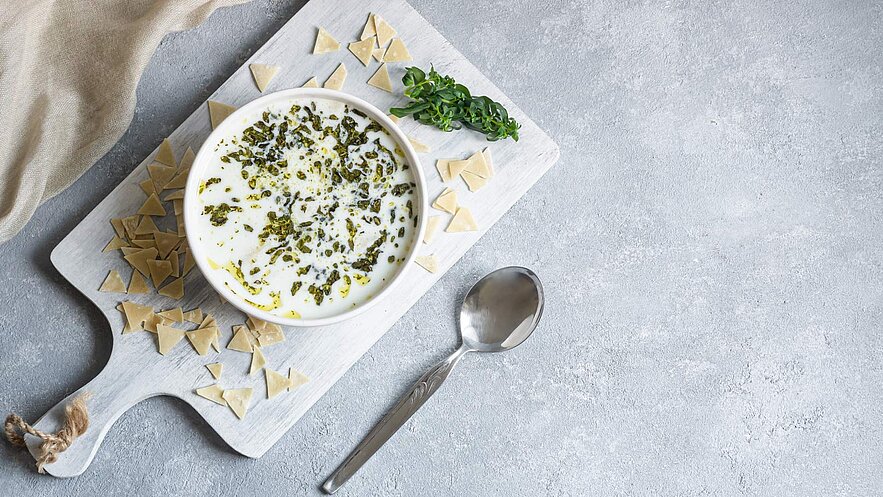 Eine Schüssel mit einer cremigen Suppe, garniert mit grünen Kräutern, steht auf einem Holzbrett. Rund um die Schüssel sind kleine Teigstücke angeordnet, und ein silberner Löffel liegt daneben. Der Hintergrund ist ein neutraler Grauton.