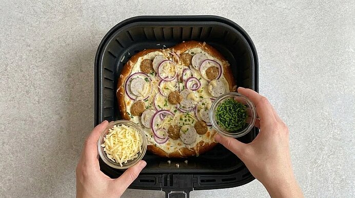 Zwei Hände halten Glasschälchen mit Käse und Schnittlauch über die belegte Brezel-Pizza im Fritteusenkorb.