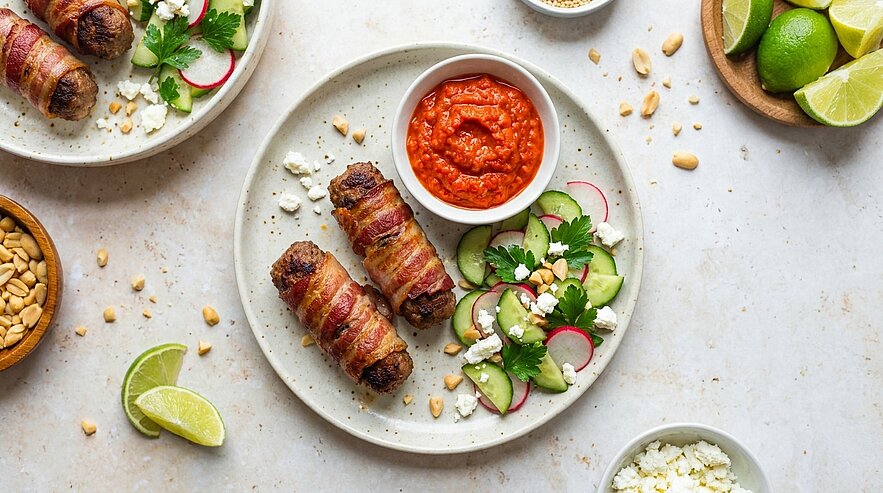 Zwei mit Speck umwickelte Fleischröllchen auf einem Teller, daneben eine Schale mit roter Dip‑Sauce und ein Salat aus Gurkenscheiben und Radieschen, Koriander, zerbröseltem Käse sowie Limettenscheibe und verstreuten Erdnüssen.
