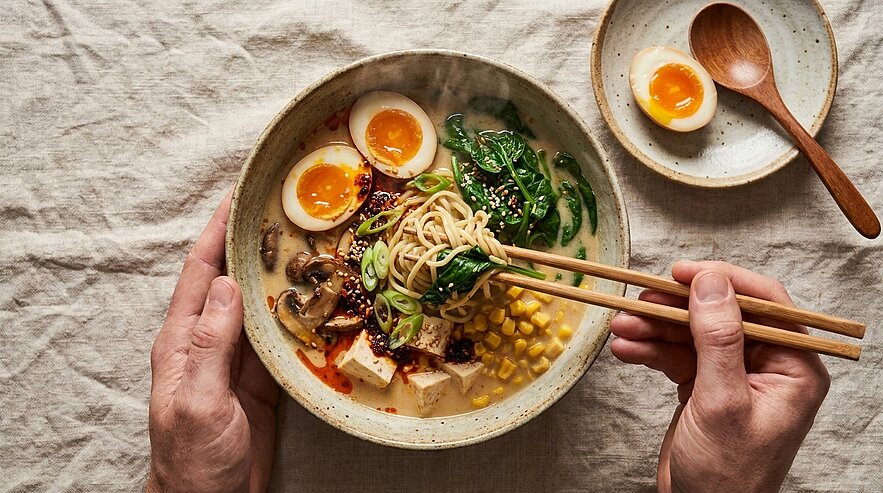Schale mit Ramen: heiße Brühe, Nudeln, weich gekochte Eier, Tofu, Spinat, Pilze, Mais und Sesam; Hände halten die Schale, Stäbchen heben Nudeln.