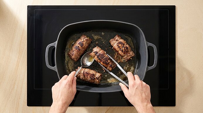 Zwei Hände halten Löffel und Zange und begießen vier gebräunte Rouladen in einer Bratform mit Bratensaft auf dem Kochfeld.
