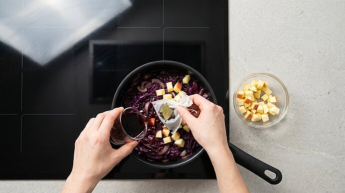 Zwei Hände fügen einem Topf mit Rotkohl eine dunkle Flüssigkeit, ein Lorbeerblatt und gewürfelte Äpfel hinzu; neben dem Topf steht eine Schüssel mit Apfelwürfeln.