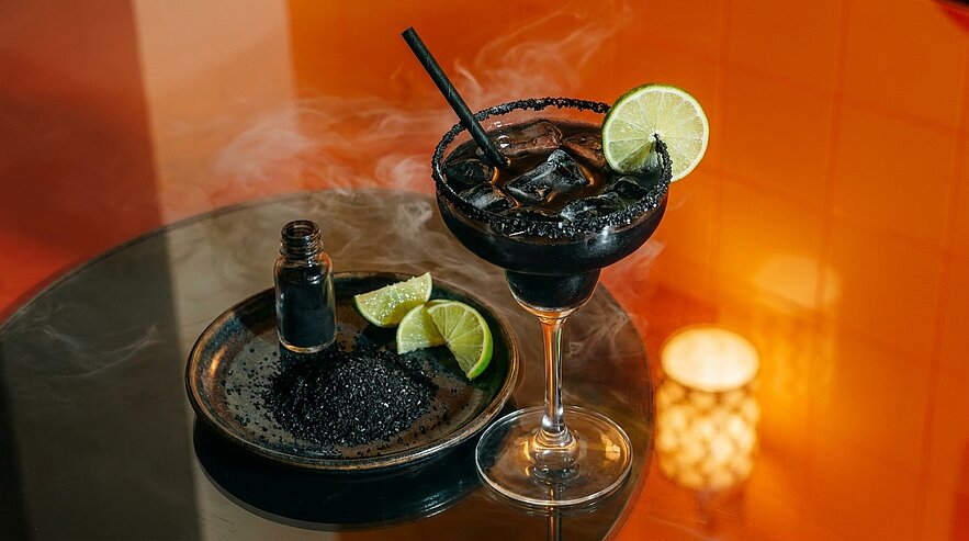 Rauchender schwarzer Cocktail in Coupéglas mit Limettenscheibe und Strohhalm, daneben Teller mit schwarzem Salz und Limettenstücken.