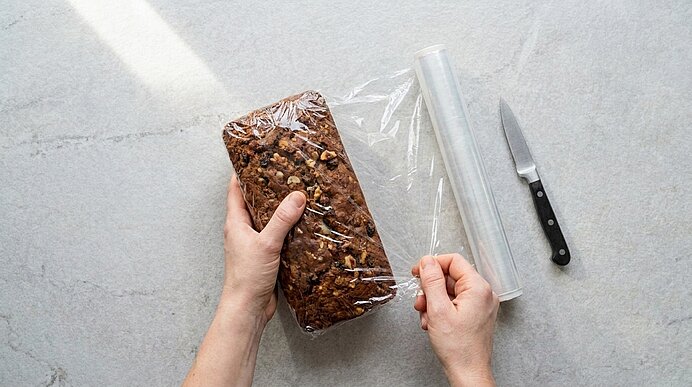 Hände verpacken ein Nussbrot in Frischhaltefolie; eine Rolle Frischhaltefolie und ein Messer liegen daneben auf hellem Untergrund.