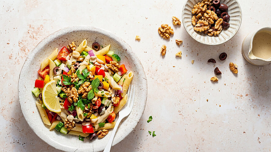 Eine Schüssel mit buntem Nudelsalat, gemischt mit Paprika, Gurke, Zwiebeln, Oliven und Walnüssen, garniert mit einem Zitronenschnitz und frischen Kräutern. Neben der Schüssel befindet sich eine kleine Kanne mit einer Sauce und eine weitere Schale mit Walnüssen.