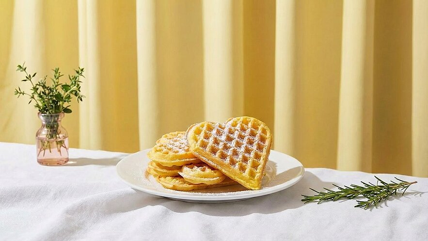 Auf einem Teller liegen mehrere Waffeln, darunter eine herzförmige Waffel, die mit Puderzucker bestäubt ist. Im Hintergrund steht eine kleine Vase mit frischen Kräutern auf einem weißen Tischcloth. Der Hintergrund ist gelb und sorgt für eine warme Atmosphäre.