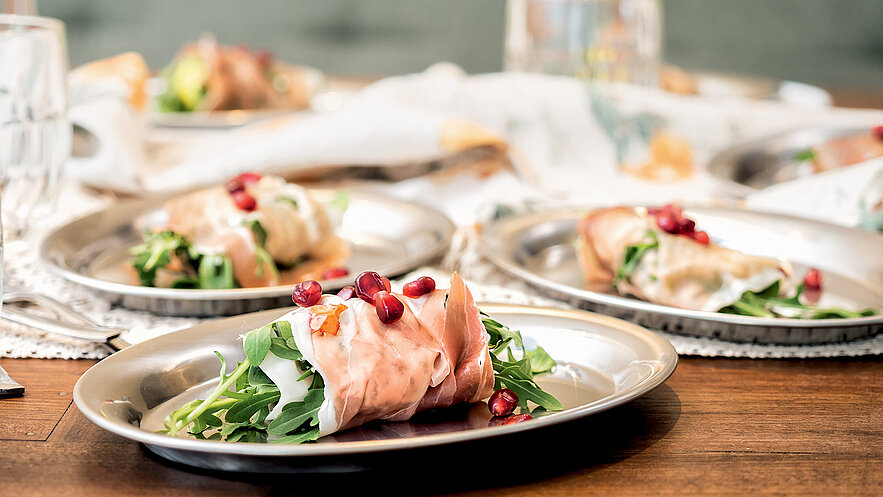 Auf dem Bild sind mehrere metallene Teller mit einer Portion Rucola-Salat und dekorativem Schinken zu sehen. Die Speisen sind mit Granatapfelkernen garniert und auf einem Holztisch angerichtet. Der Hintergrund zeigt weitere Teller und eine Tischdecke.