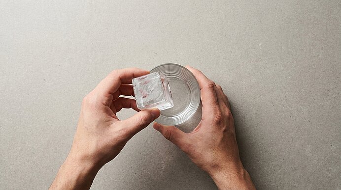 Zwei Hände, eine setzt einen großen Eiswürfel in ein klares Glas mit Wasser.