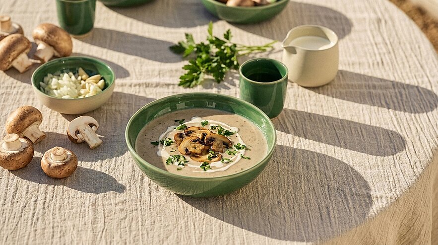 Schale mit cremiger Champignon‑Suppe, garniert mit gebratenen Pilzscheiben und Petersilie; daneben rohe Champignons, gehackte Zwiebeln/Knoblauch und ein Sahnekännchen.