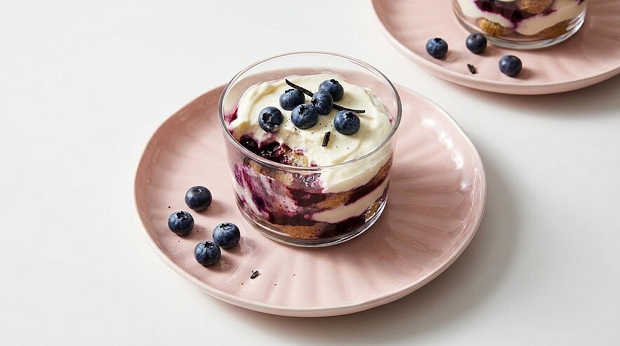 Glas mit geschichtetem Dessert aus heller Creme und Blaubeerkompott, oben mit frischen Blaubeeren garniert, auf rosa Untertasse platziert.