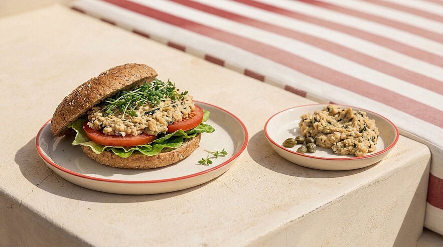 Sandwich aus Vollkornbrötchen mit Salatblatt, Tomatenscheiben, cremigem Aufstrich und Sprossen; daneben kleine Schale mit Aufstrich und Kapern.