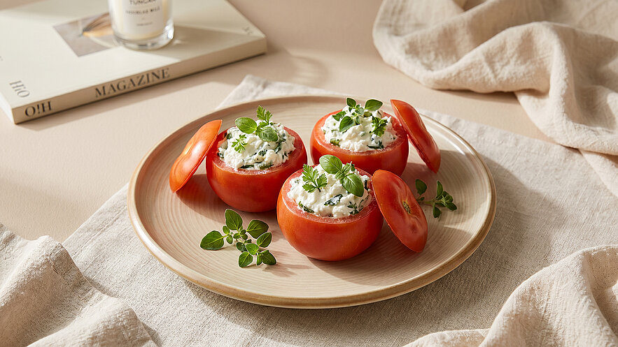 Auf einem dekorativen Teller liegen drei halbierte Tomaten, gefüllt mit einer herzhaften Creme und garniert mit frischem Oregano. Im Hintergrund sind unscharf ein Magazin und eine gedeckte Oberfläche zu sehen.