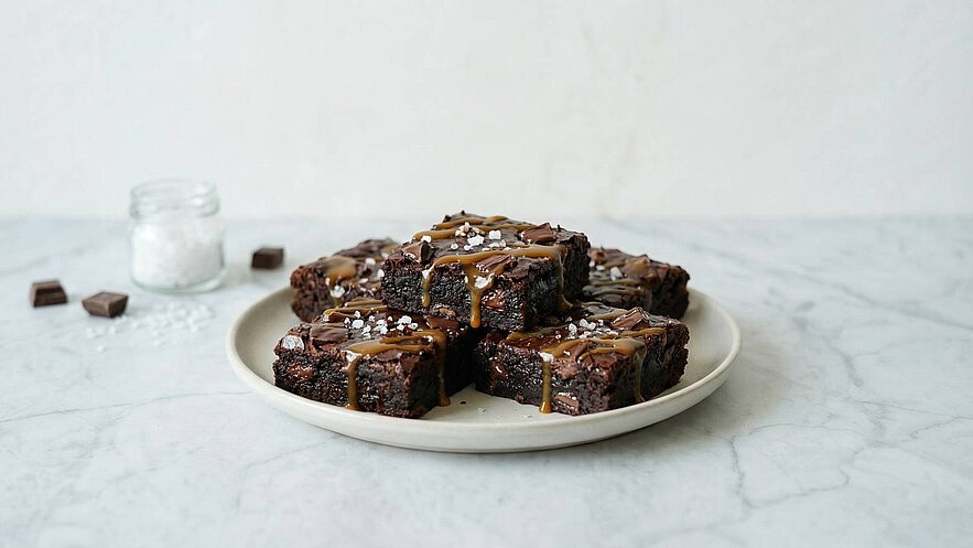 Ein weißer Teller mit mehreren Stücken schokoladiger Brownies, die mit Karamell und einer Prise Salz garniert sind. Die Brownies liegen auf einem marmorierten Untergrund. Neben dem Teller steht ein Glas mit Zucker.