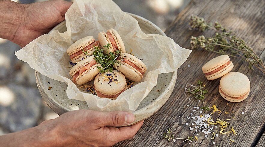 Zwei Hände halten eine Schale mit Pergamentpapier und mehreren hellen Macarons mit rosa Füllung, garniert mit Kräutern und Blüten; auf dem Holzuntergrund liegen weitere Macarons, Kräuter und grobes Salz.