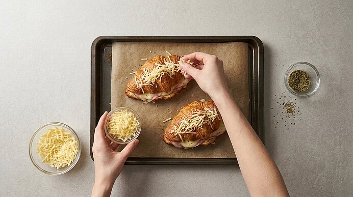 Zwei Hände bestreuen gefüllte Croissants auf einem Backblech mit geriebenem Käse.